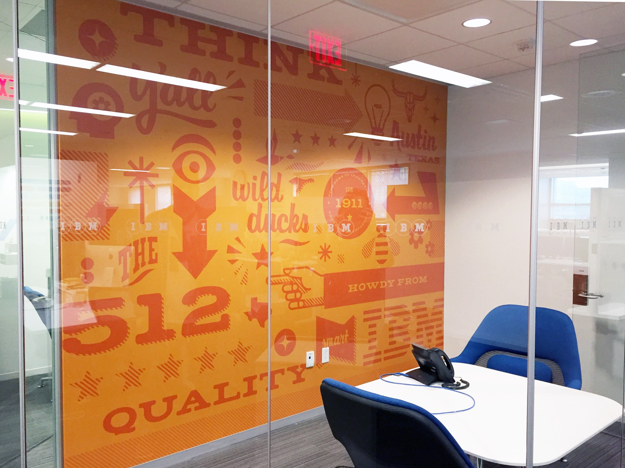 Orange and red-orange typographic mural behind a glass conference room wall with words like Think, Y'all, wild ducks, The 512, Quality and Austin Texas