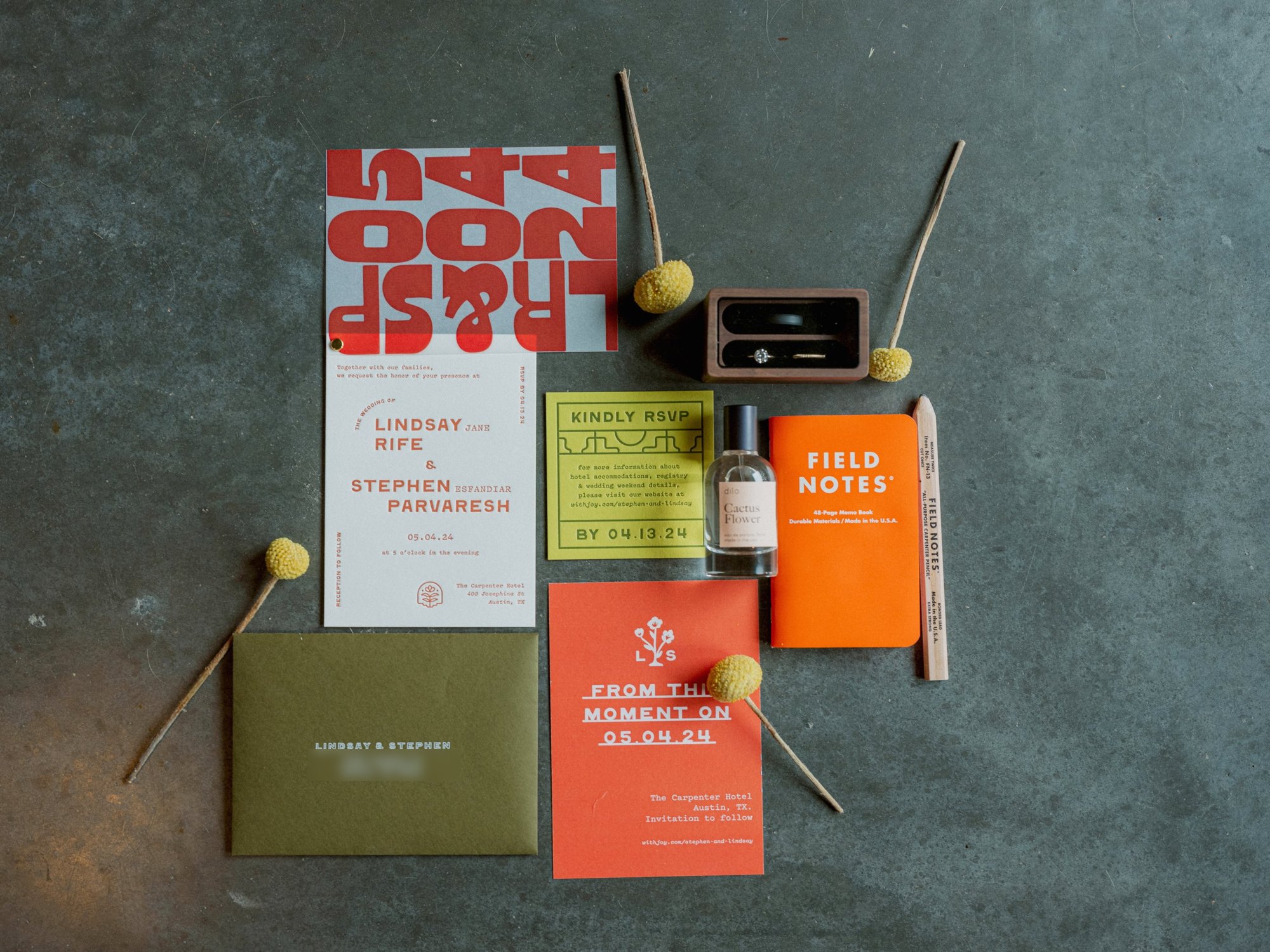 Flat lay of the printed suite on concrete: vellum numeral wrap over the letterpress invitation, chartreuse RSVP card, orange save the date and olive envelope, with rings and billy button flowers
